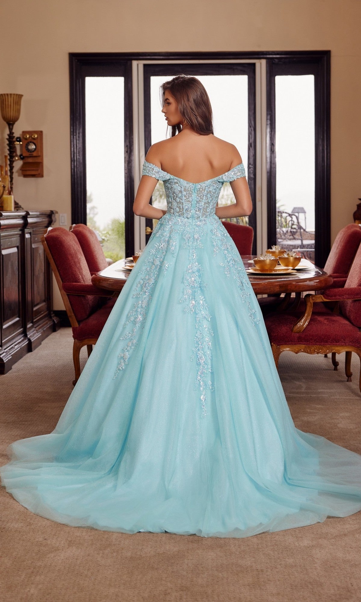 A woman in the Nox Anabel H1881 long prom dress—a light blue off-the-shoulder tulle gown with floral details—stands with her back to the camera in a formal dining room.